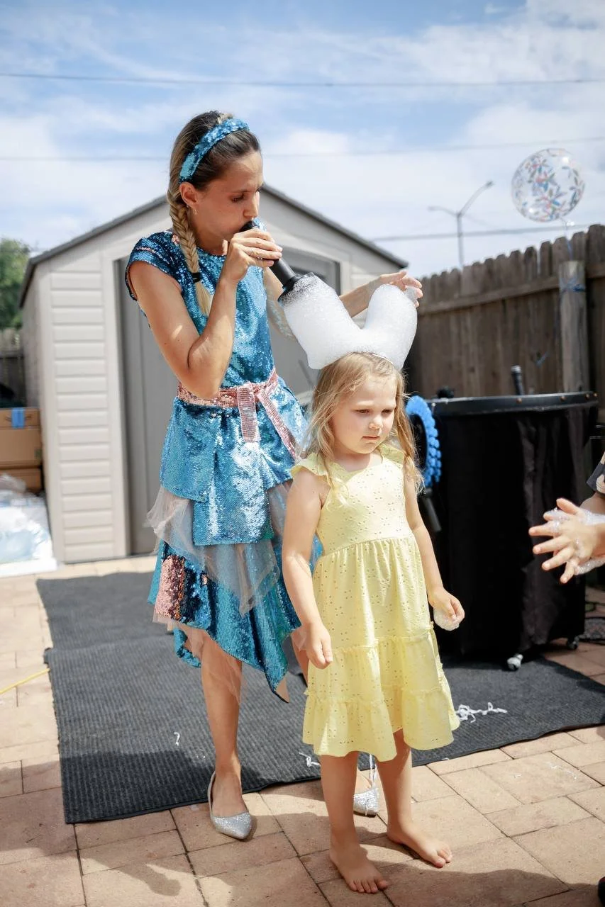 Woman dressed as a fairy godmother blowing bubbles over a young girl in a yellow dress at a backyard birthday party.