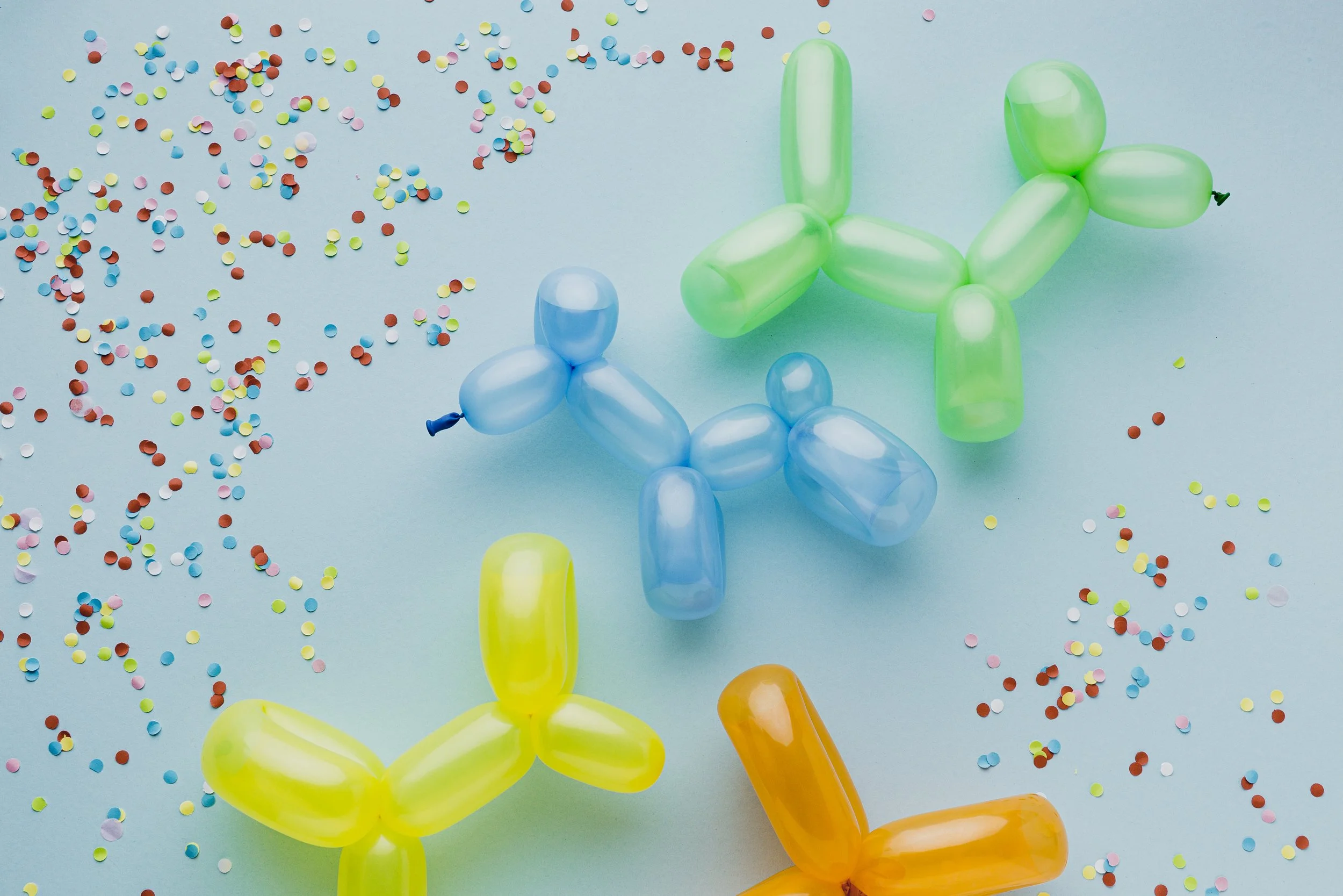 Colorful balloon animals, including a blue dog, a yellow dog, and a green dog, on a light blue background with scattered multicolored confetti.