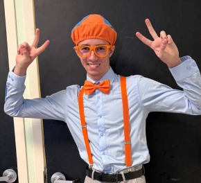 Person dressed as a chef wearing an orange hat, orange suspenders, glasses, and a bow tie, making peace signs with both hands in front of a black chalkboard.