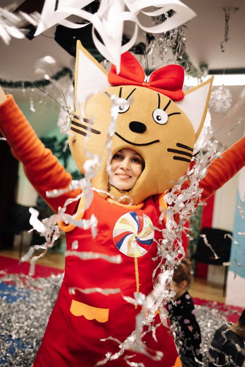 Person in a festive costume with a large cartoon cat face headpiece, red bow, and red outfit, celebrating with confetti and falling silver streamers.