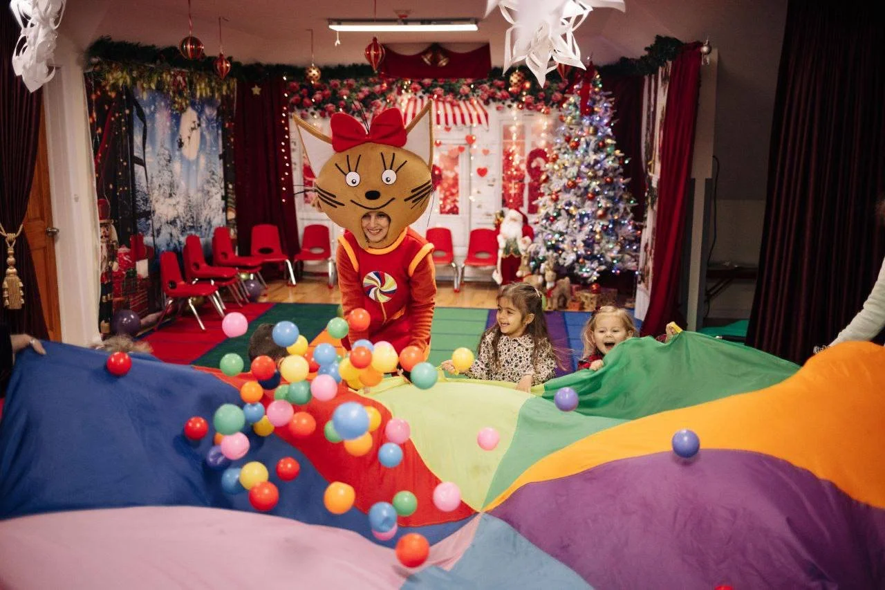 Children playing with a colorful parachute with small balls inside. A person in a large cat costume with a red bow on its head is in the center. The background features a decorated Christmas tree, holiday garlands, and a festive scene with red curtains and holiday ornaments.