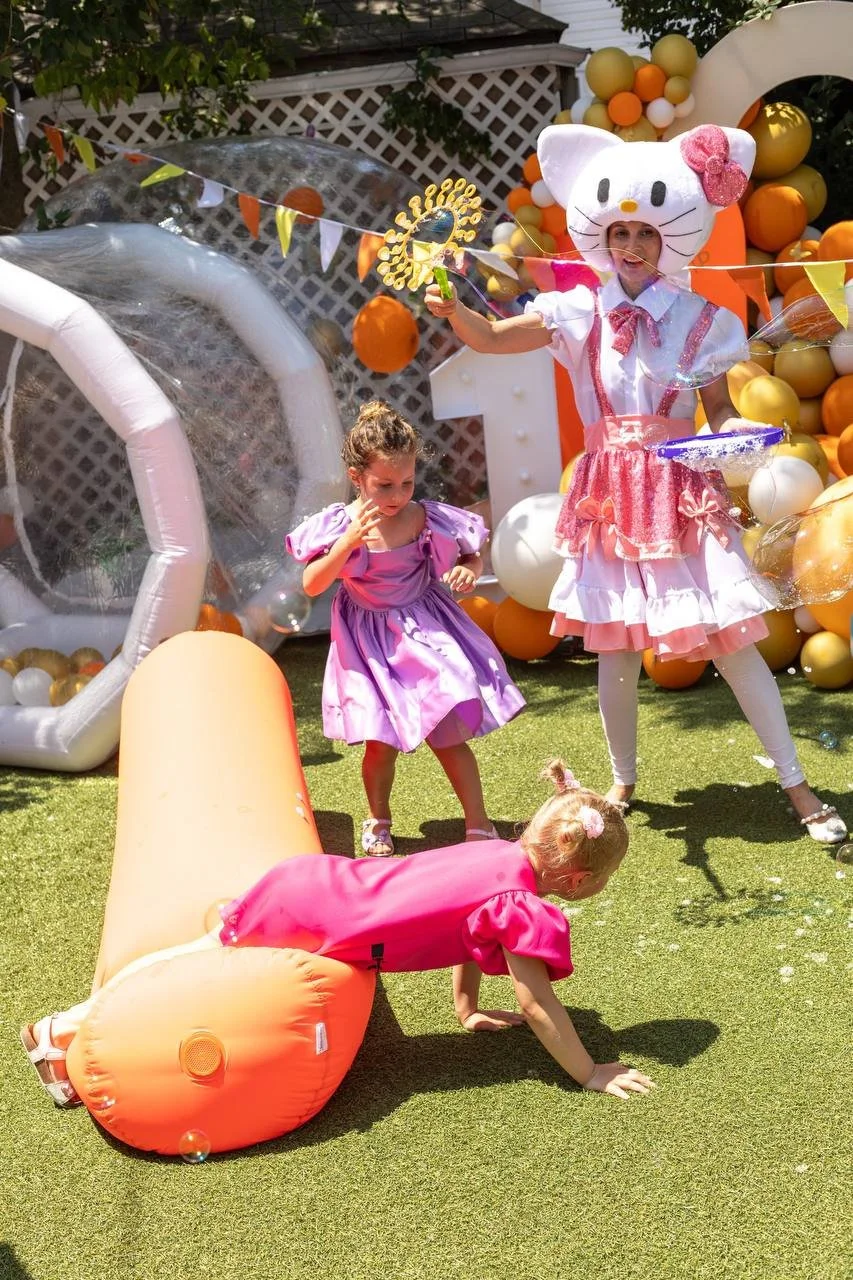 A children's birthday party outdoors with a Hello Kitty themed entertainer in costume and children playing on the grass near balloons and decorations.