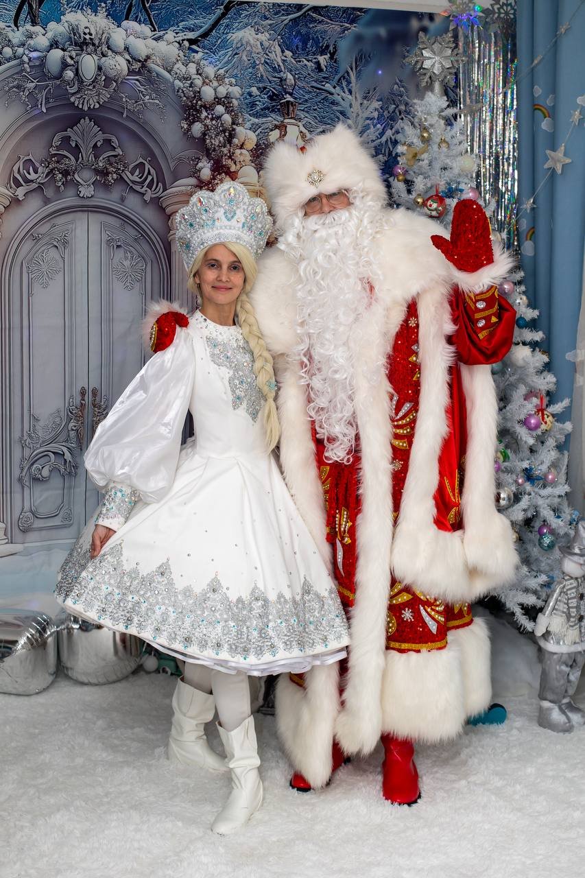 A woman dressed as the Snow Queen and a man dressed as Santa Claus standing together in a festive, snowy winter scene. The Snow Queen is wearing a white dress with silver embroidery and a tall, ornate crown, while Santa Claus is wearing a traditional red and white outfit with a fur hat and mittens. There are decorated Christmas trees and winter-themed decorations in the background.