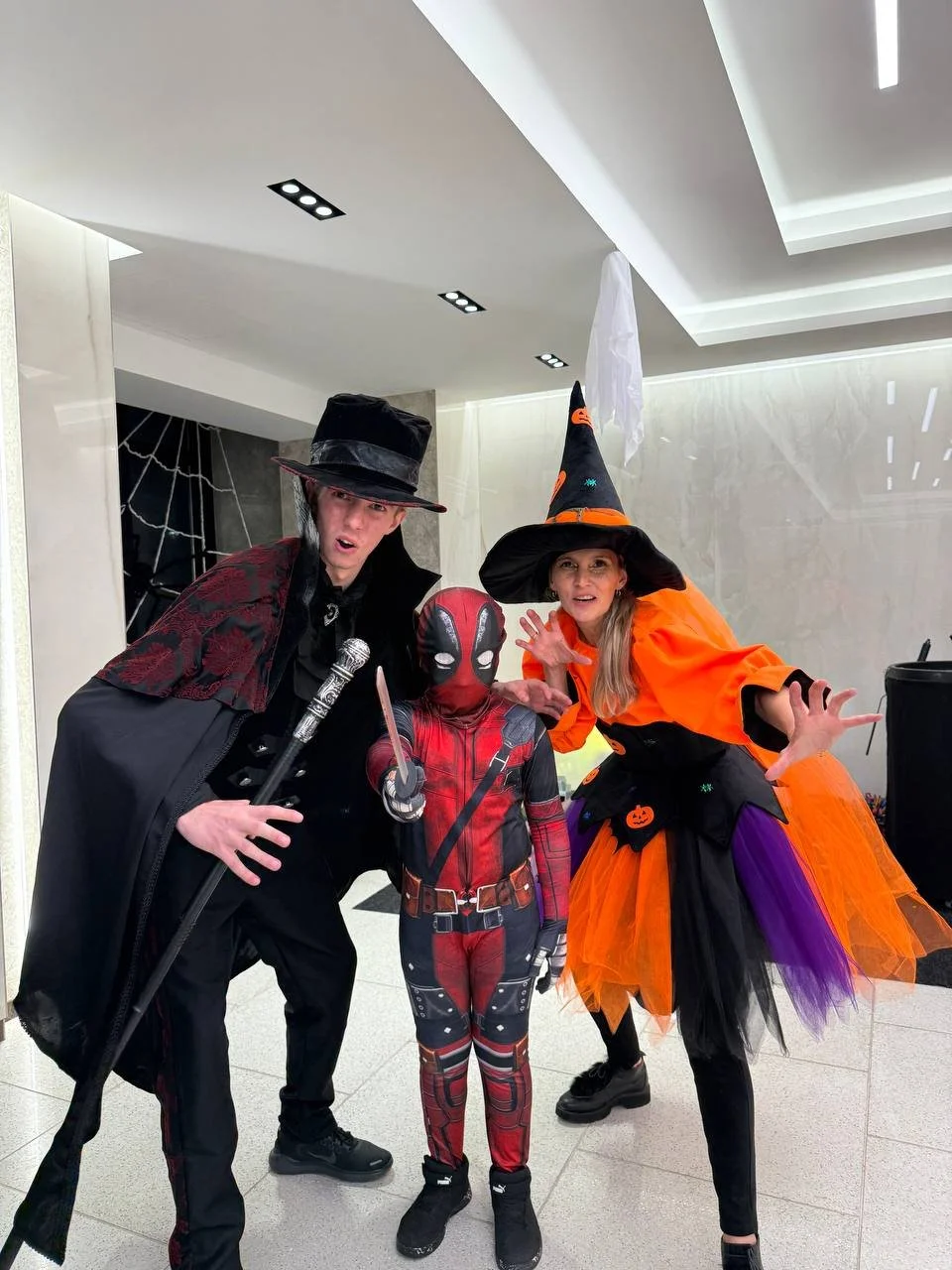 Three people dressed in Halloween costumes: a young boy as Deadpool, a woman as a witch, and a man as a wizard, posing indoors.