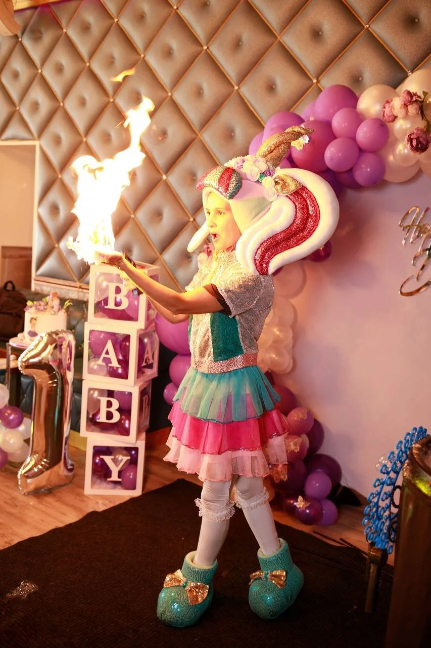 A young girl in colorful, whimsical costume holding a flaming object at a birthday party with balloons and decorations, including a large number '1' balloon and blocks spelling 'BABY'.