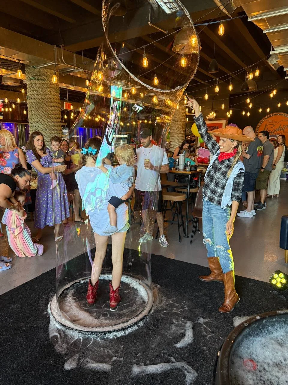 A woman in Western-style clothing and a large cowboy hat creates a giant soap bubble around a young girl inside a restaurant or bar with an industrial decor. Several people, mostly women and children, watch and enjoy the event, with string lights hanging from the ceiling adding to the lively atmosphere.