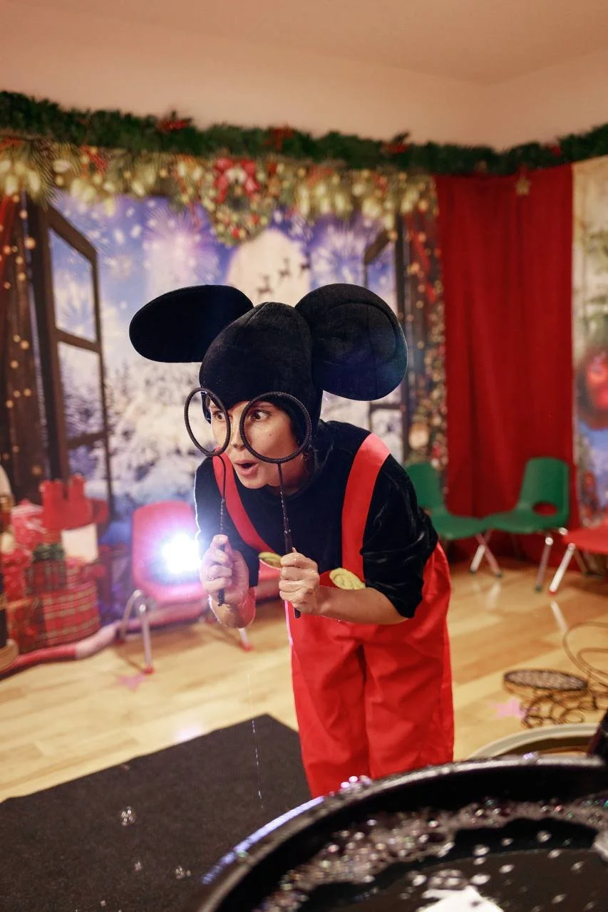 Person dressed up as Mickey Mouse holding a magnifying glass in a festive room decorated for Christmas, with a window showing a snowy outdoor scene and Christmas ornaments and gifts in the background.