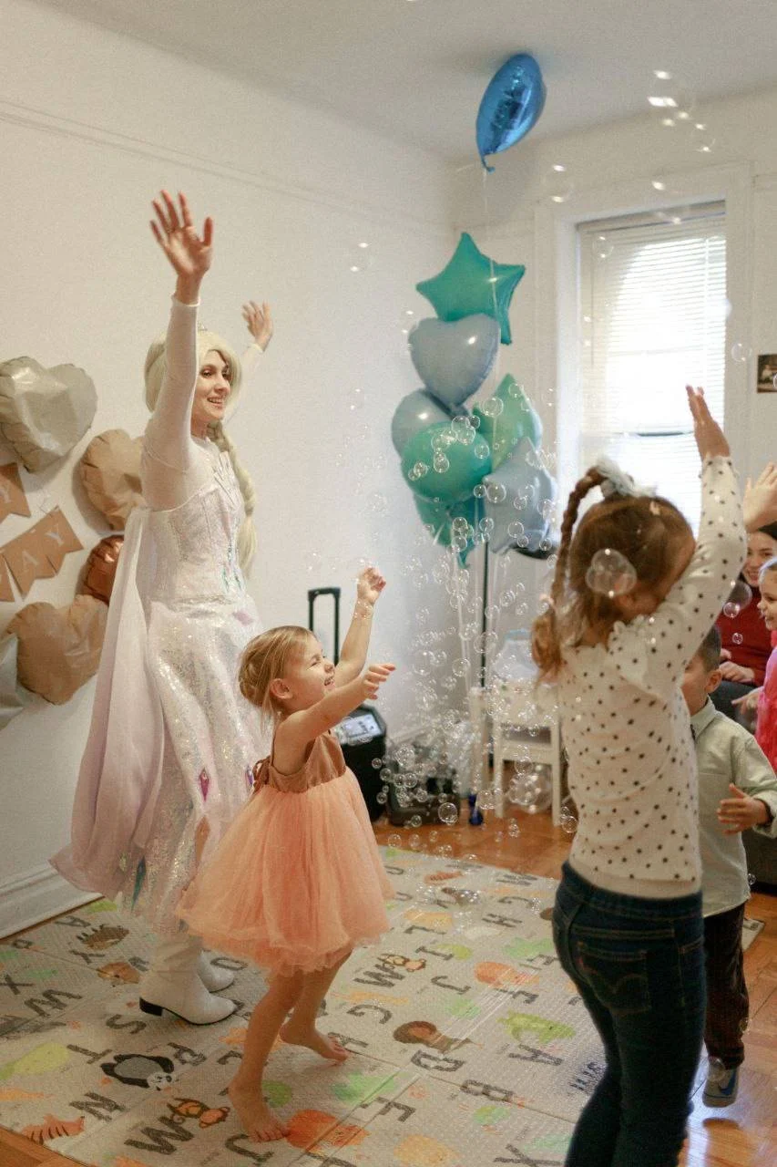 Children and a woman dressed as Elsa from Frozen are playing with bubbles at a party, with balloons and decorations in the background.