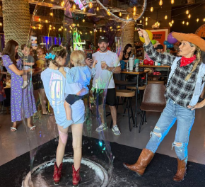 Kids and adults celebrating at a party with balloons, confetti, and string lights, dancing and having fun inside a decorated venue.