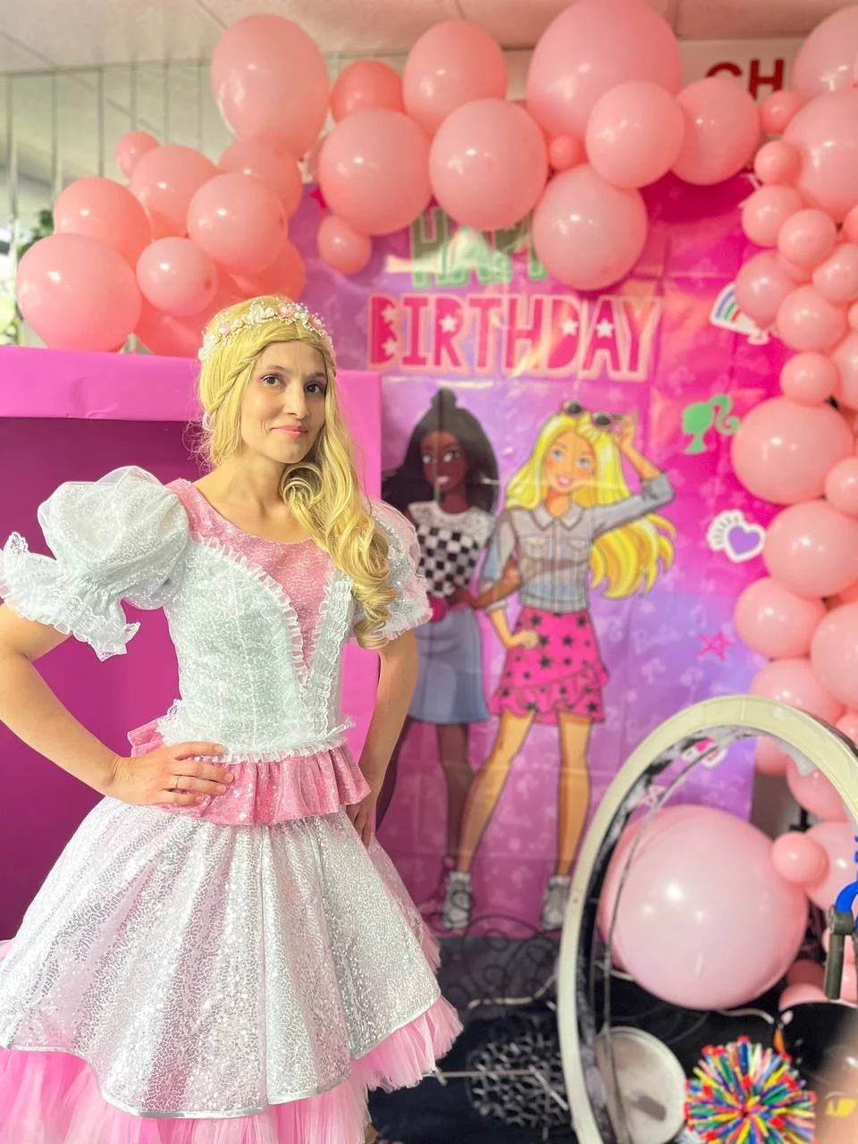 A woman dressed as a princess posing at a birthday party decorated with pink balloons and a colorful backdrop featuring cartoon girls, with one hand on her hip and smiling slightly.