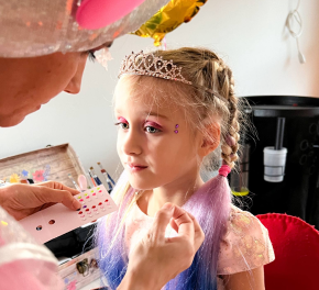 A young girl with colorful makeup and a tiara getting her face painted by an adult.