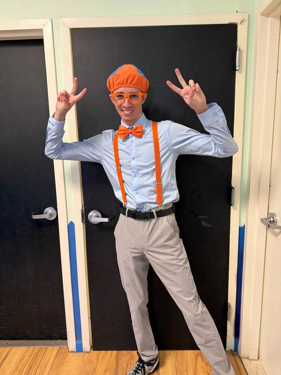 Person dressed in colorful costume with orange suspenders, bow tie, glasses, and a cap, standing in front of blackboards, making a peace sign with both hands, smiling.