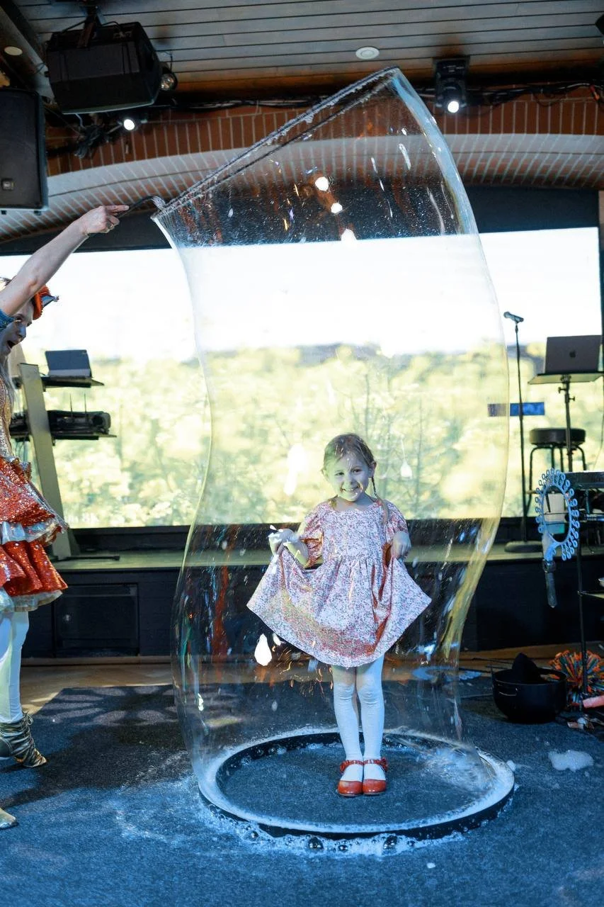 A young girl in a floral dress, white tights, and red shoes standing inside a large soap bubble with a person holding a wand to create the bubble. The girl is smiling. The background shows a screen with an outdoor scene and a stage setup with equipment.
