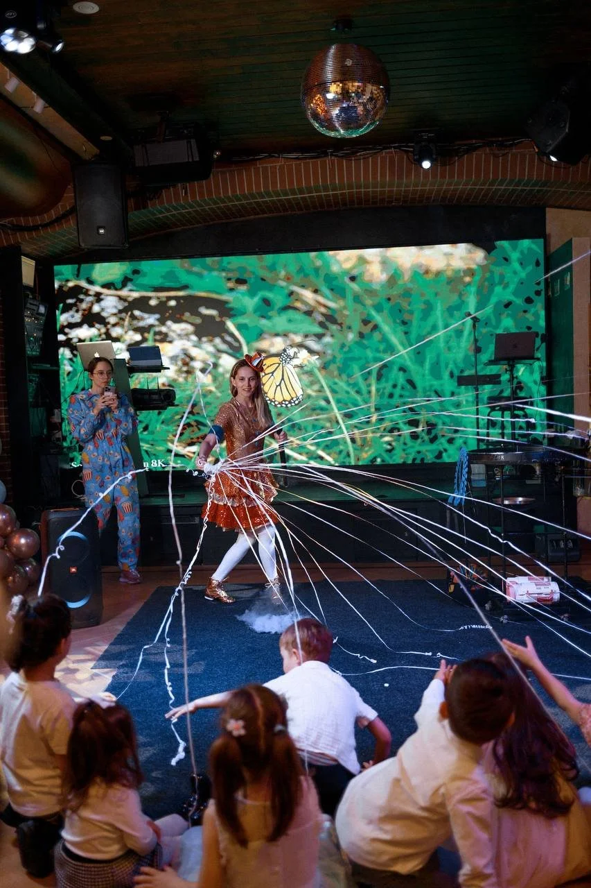 Children watching a performer with streamers and a butterfly costume on stage, in a room with a large screen showing a nature scene.
