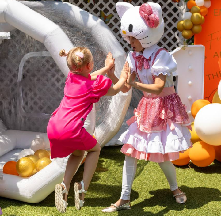 A young girl in a pink dress high-fives a person dressed as Hello Kitty, who is wearing a pink and white dress with a Hello Kitty headpiece, at an outdoor event with balloons and a large inflatable that resembles an apple in the background.