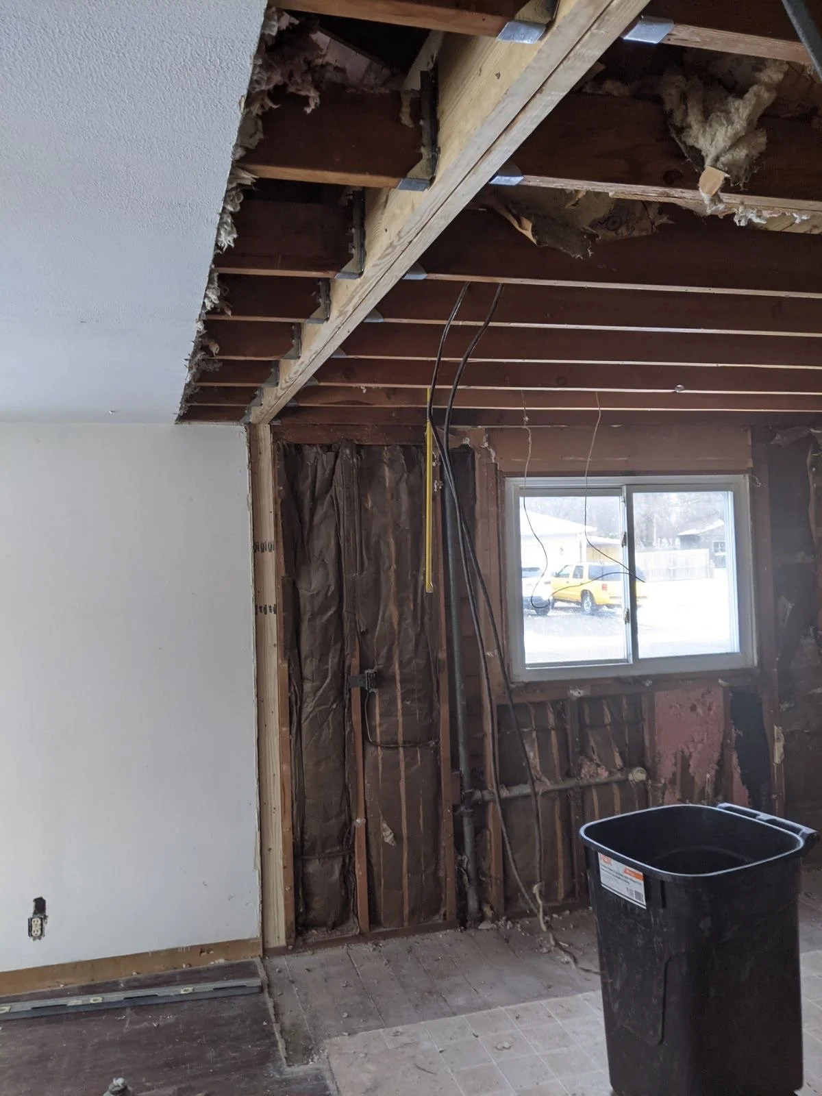 Room under renovation with exposed wall studs, partially removed drywall, a window, electrical wiring, and construction debris.