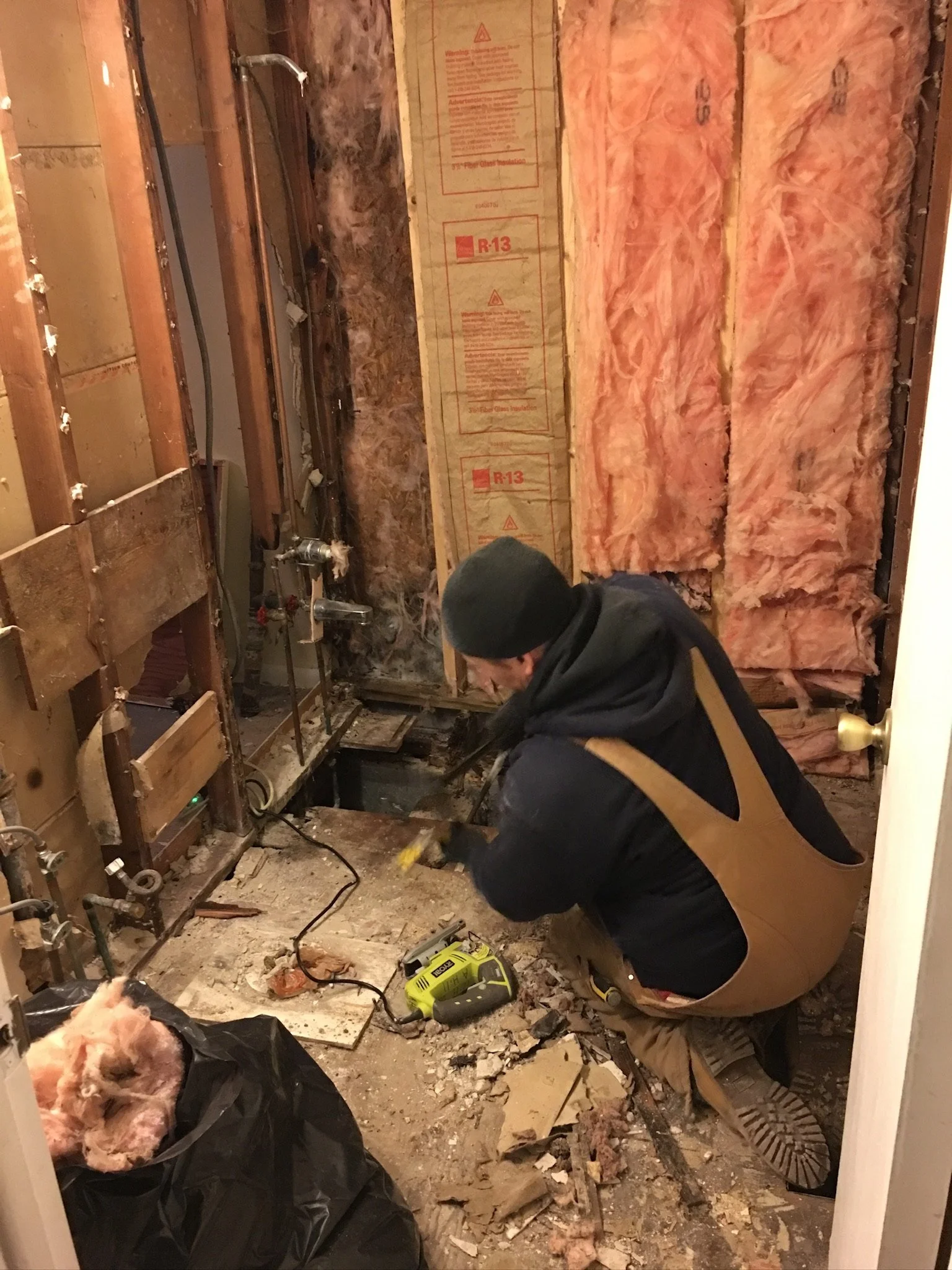 A person working on a demolition project inside a room, removing walls and insulation with construction tools on the floor, exposed pipes, insulation, and debris.