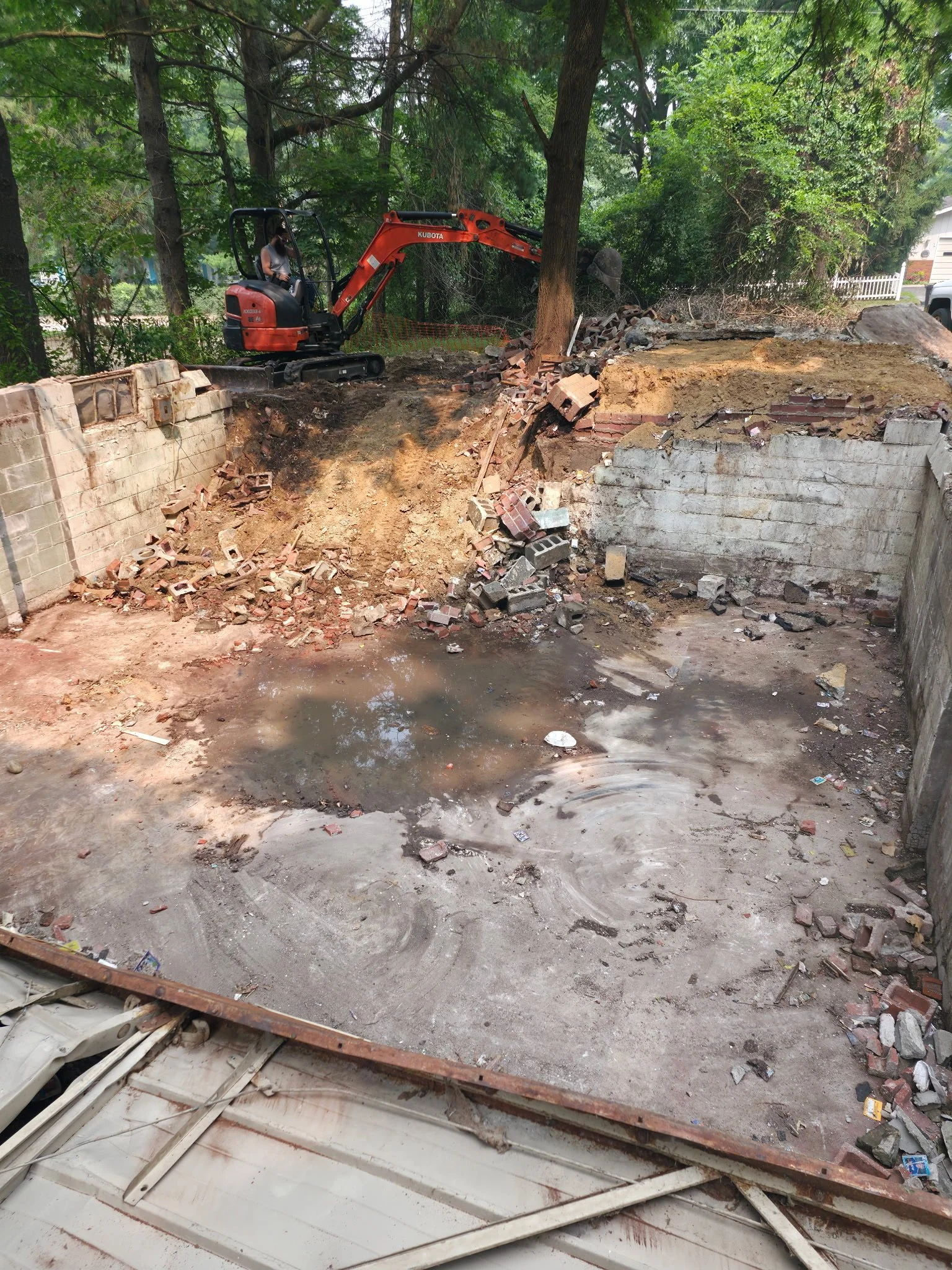 Construction site with a small orange excavator demolishing a wall, debris scattered around, in a wooded area.