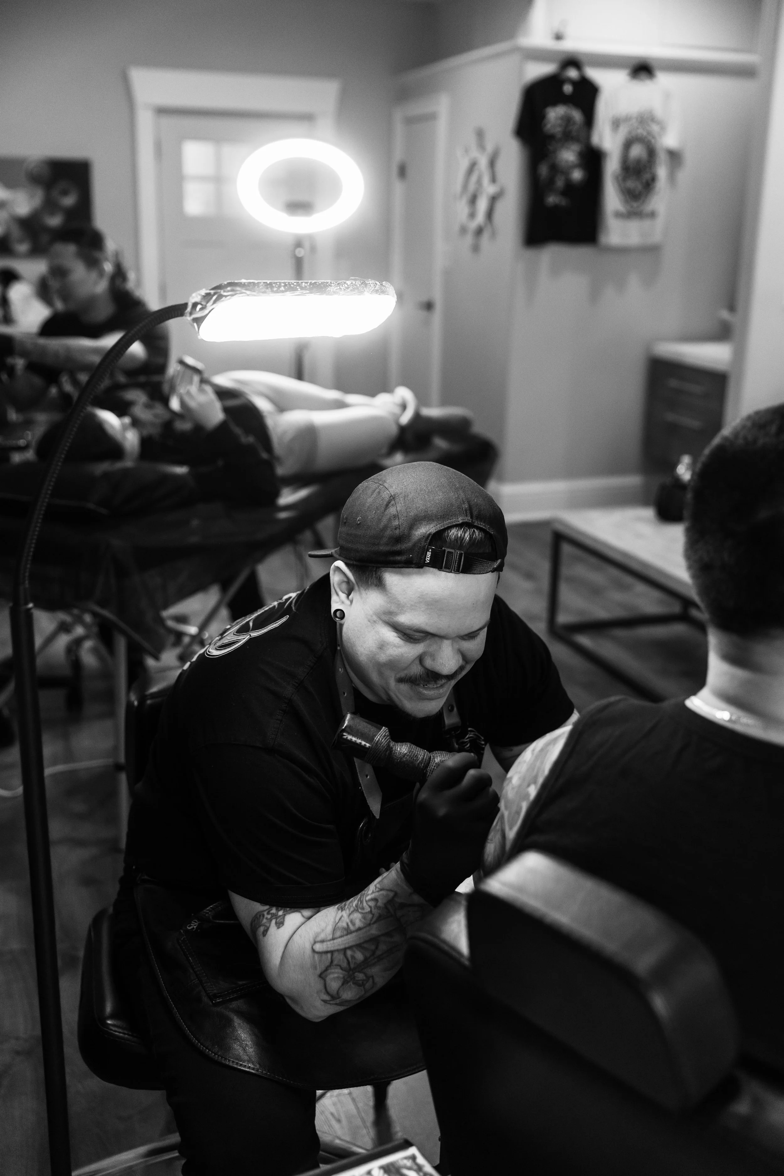 A tattoo artist with a cap, gloves, and tattoos on his arms working on a client's arm in a tattoo studio with artwork and clothing hanging on the walls.