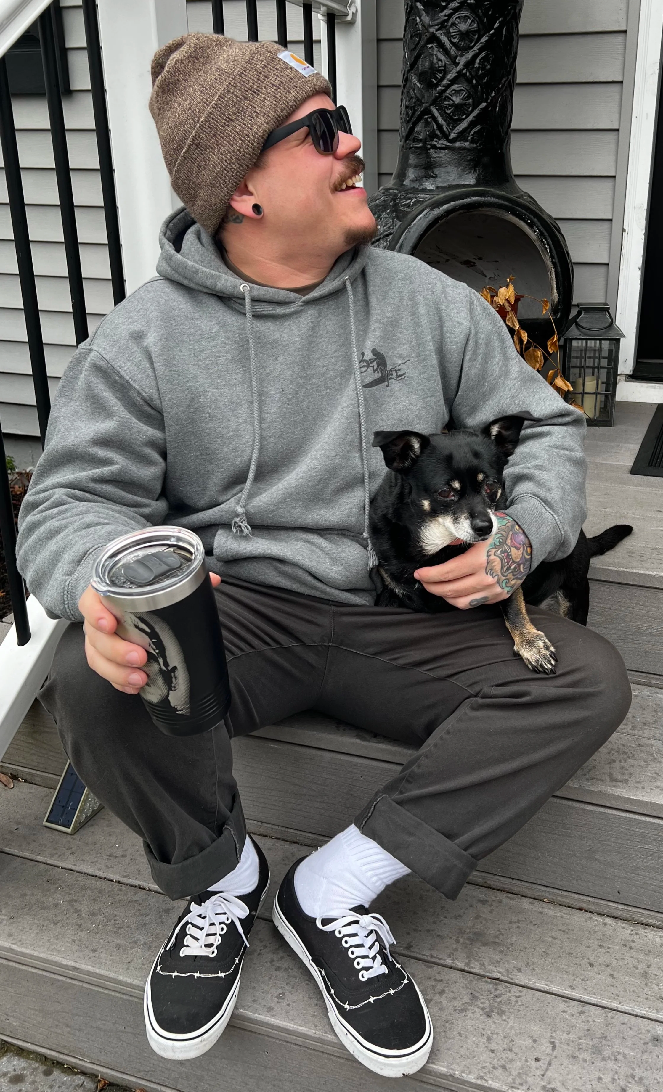 A man sitting on a wooden deck holding a small black and tan dog and a black insulated cup, wearing sunglasses, a gray hoodie, black pants, and black sneakers with white soles. There is a chimney and house siding in the background.