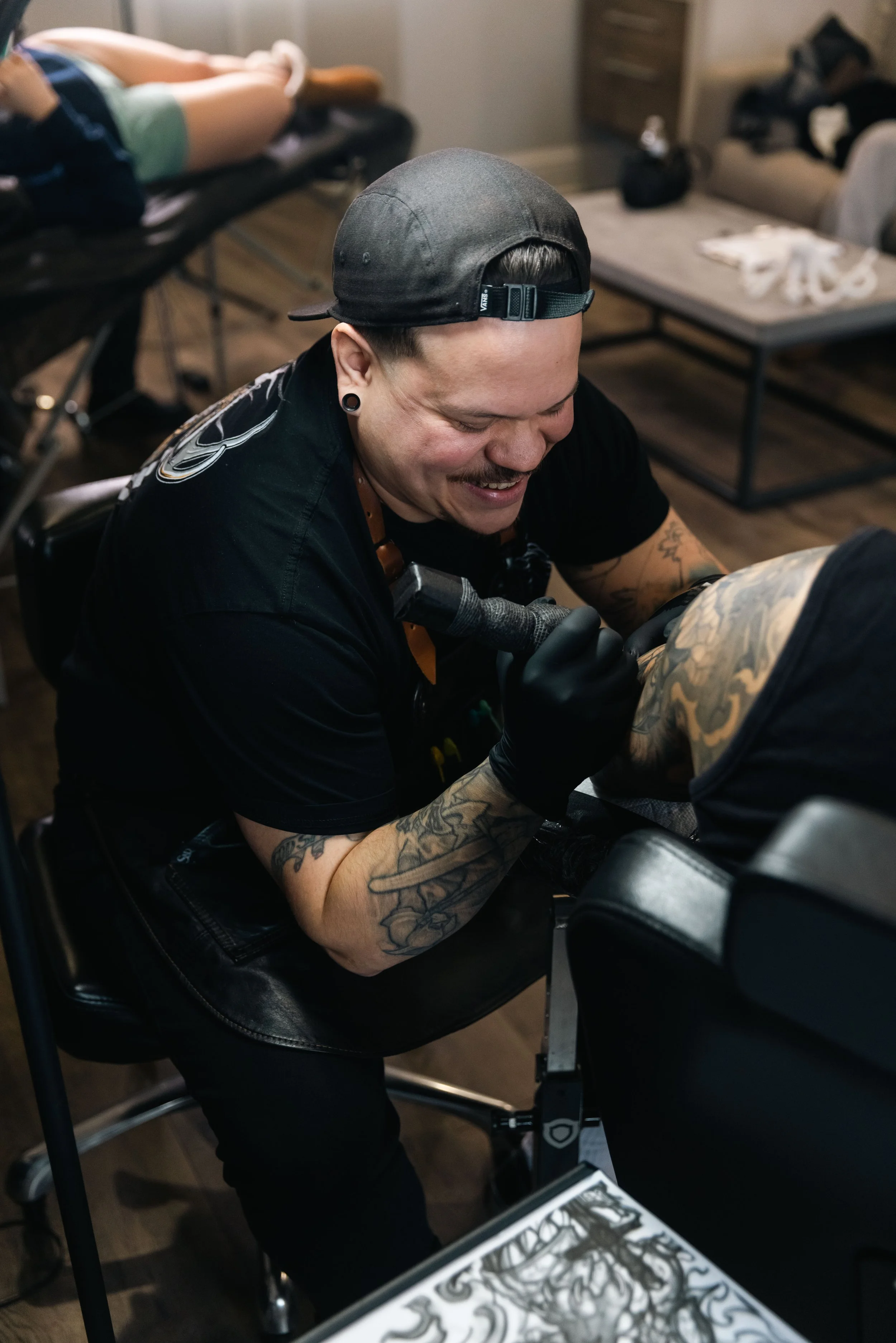 A man with tattoos, wearing a black cap backward and black gloves, smiling and working on a tattoo on another person's arm in a tattoo studio.