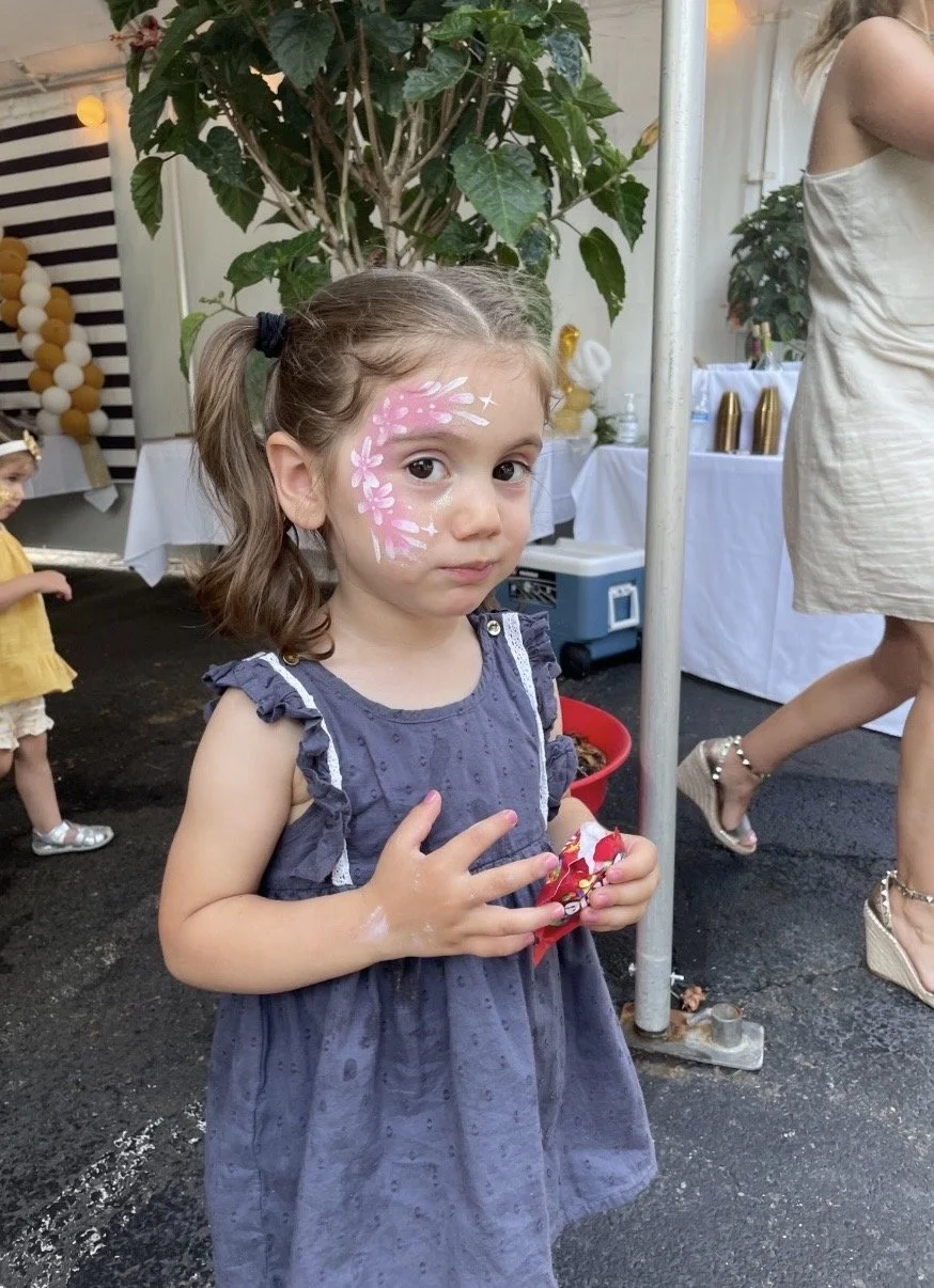 This is a photo of a young girl with face paint makeup, wearing a blue dress, standing at an outdoor event. She has brown hair in pigtails and is holding a wrapped gift. In the background, there is another child with face paint, plants, and tables wi