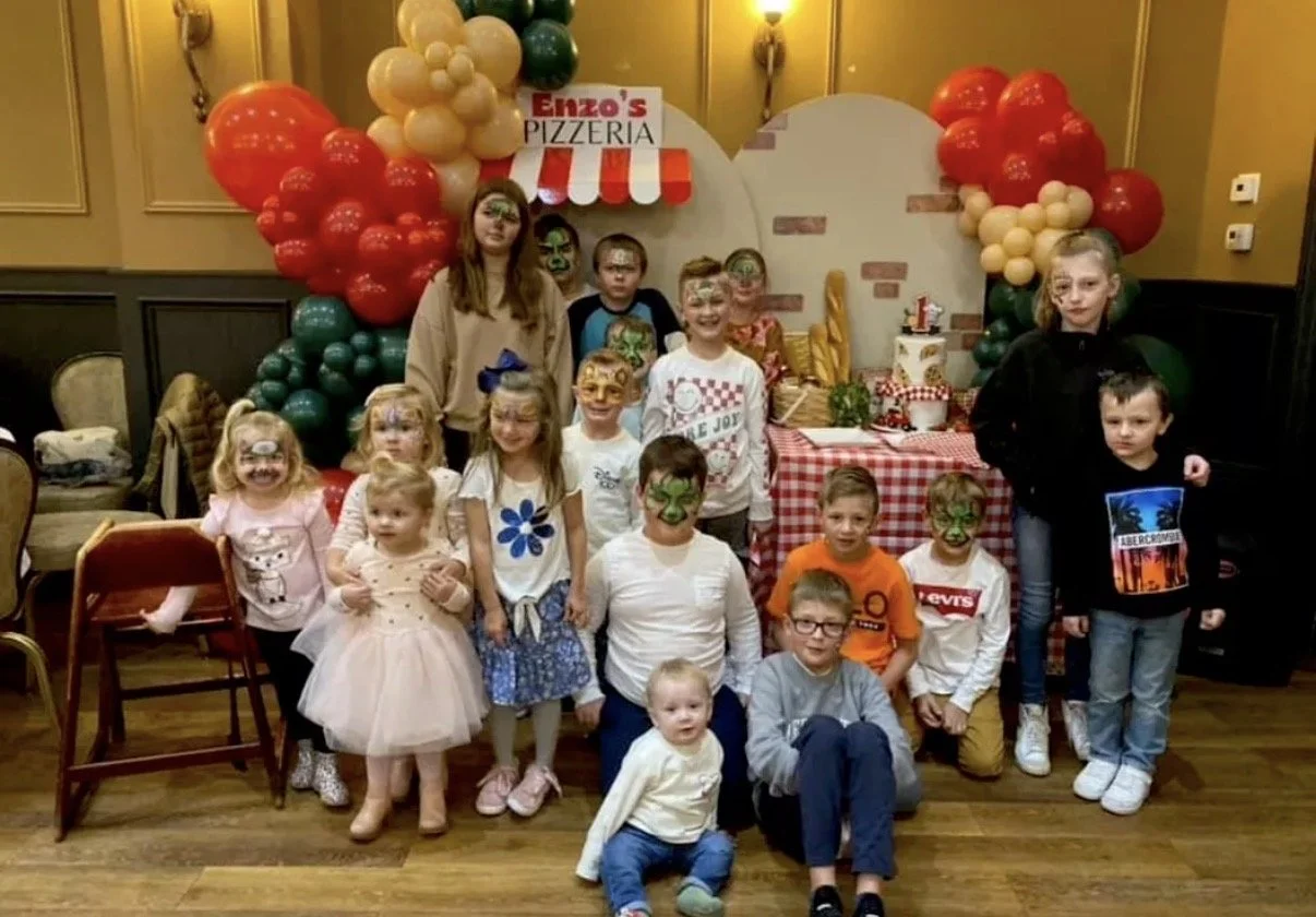 Children at a birthday party with face paint, posing in front of a decorated table and balloon arch at Enzo's pizzeria.