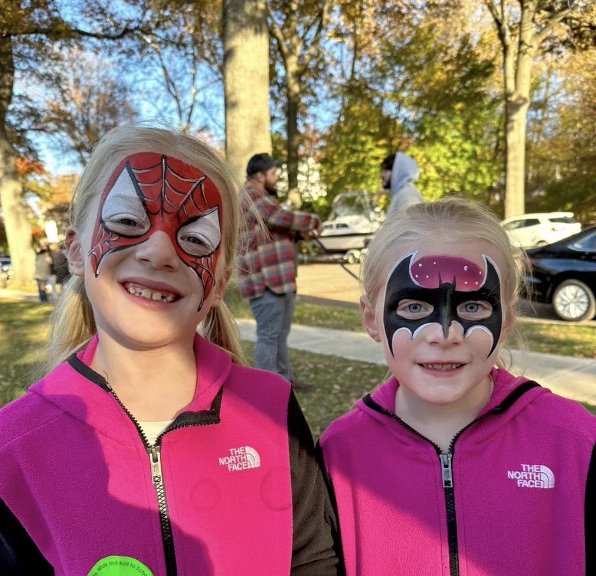 Two young girls with face paint, smiling outdoors in autumn, wearing pink The North Face jackets, with trees and parked cars in the background.