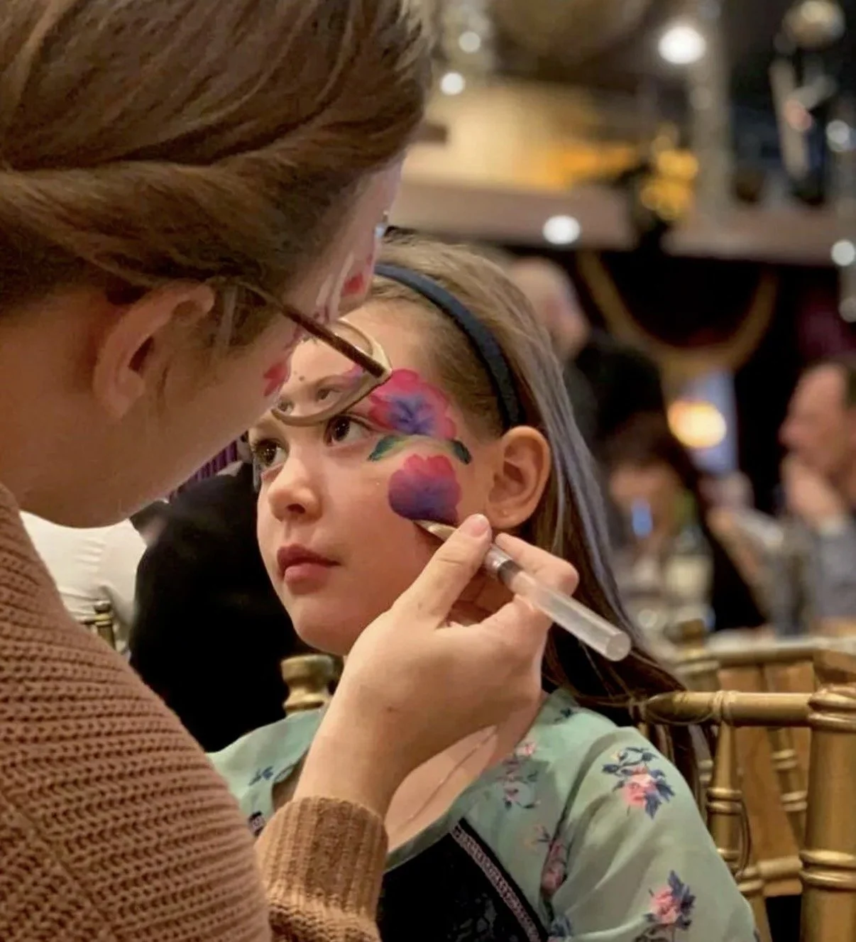 A young girl getting face painted with colorful flowers by an adult in a crowded indoor setting.