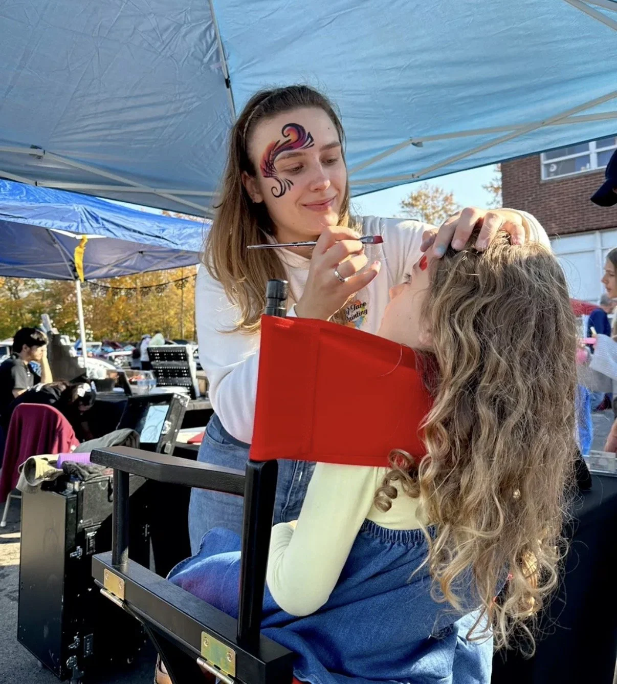 A young woman with face paint designs doing face painting on a girl sitting on a chair outdoors under a blue canopy.