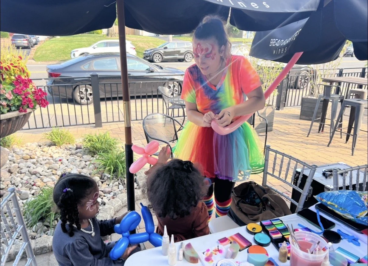 A woman dressed as a rainbow clown making balloon animals for two children at an outdoor birthday party. The children have face paint and are holding balloon animals. There are party supplies on the table and patio chairs under umbrellas.