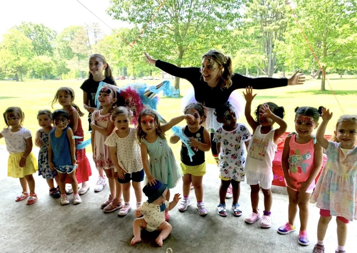 A group of young children and an adult dressed as a clown, with face paint, posing outdoors under a tent in a park with green trees.