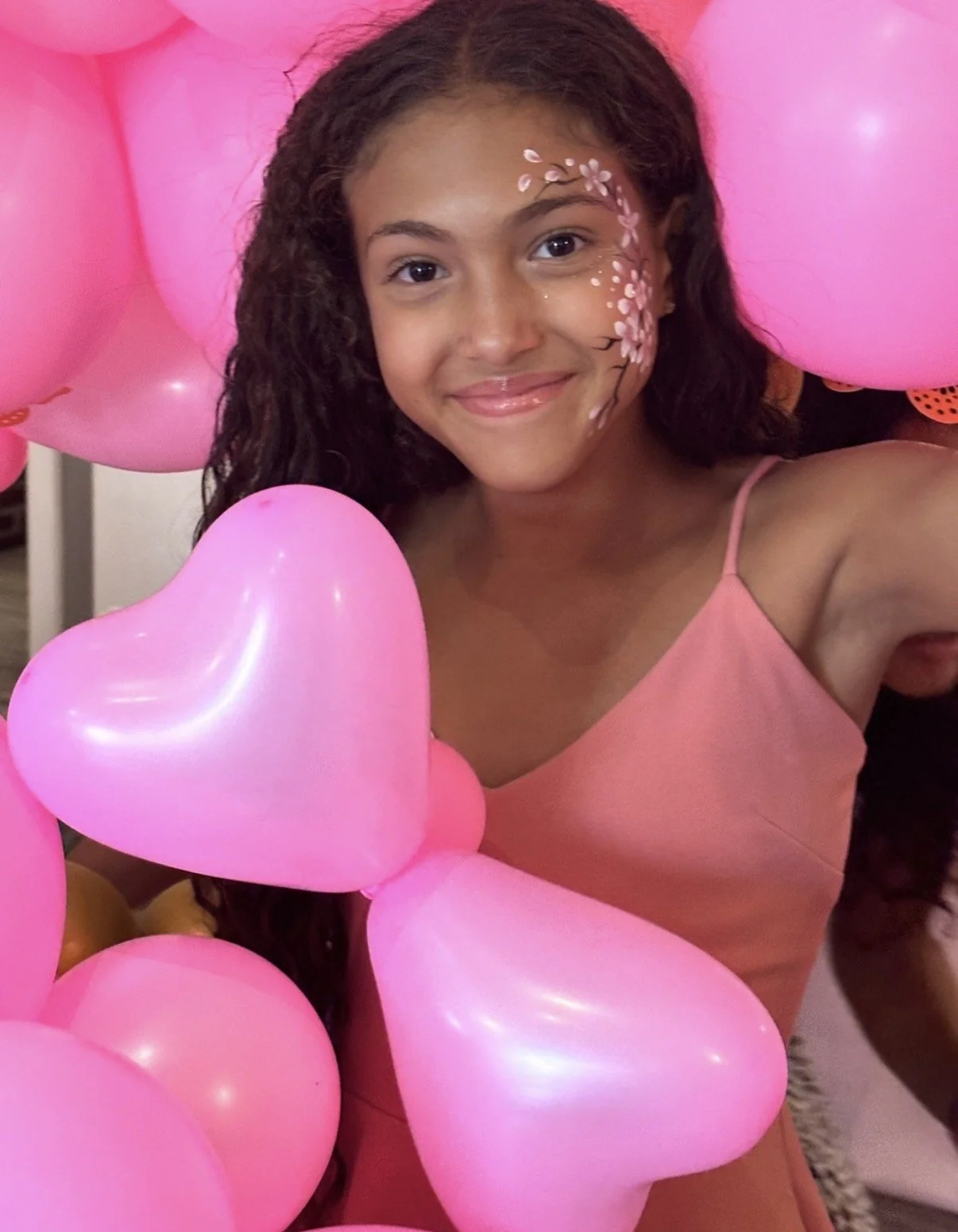 A young girl with curly hair and face paint of pink flowers and white dots, smiling, surrounded by pink heart-shaped balloons, wearing a pink dress.