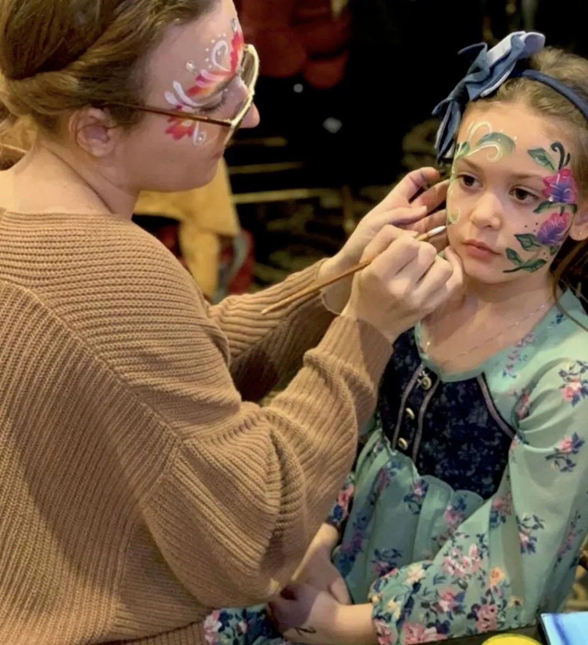 A woman doing face paint on a young girl with face paint designs and floral clothing.