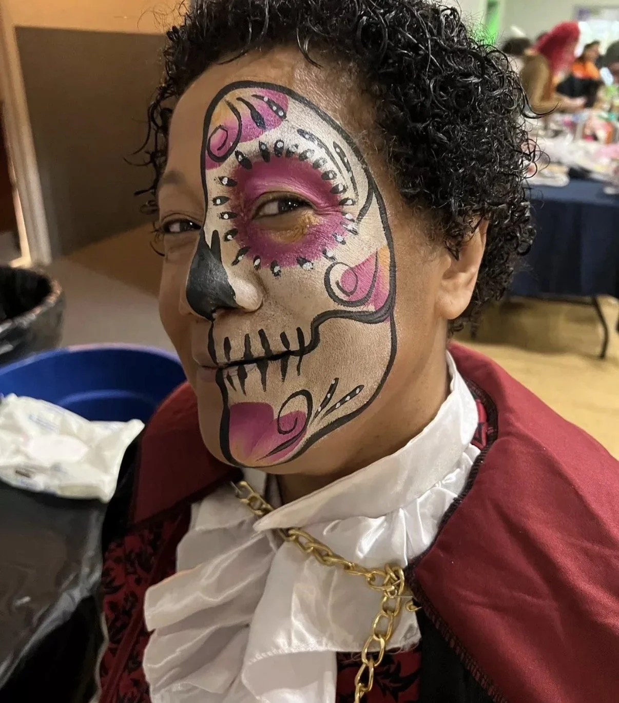 A woman with curly hair has face paint that resembles a colorful sugar skull, with pink, white, and black designs covering half her face.
