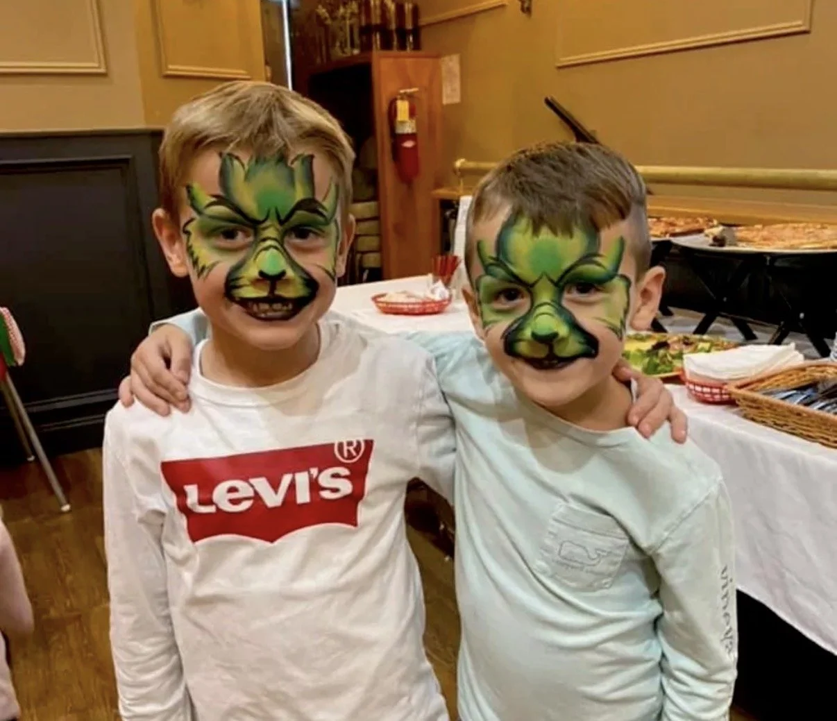 Two boys with painted faces resembling green tiger faces, smiling and posing together at a party or event, with a table and pizza in the background.
