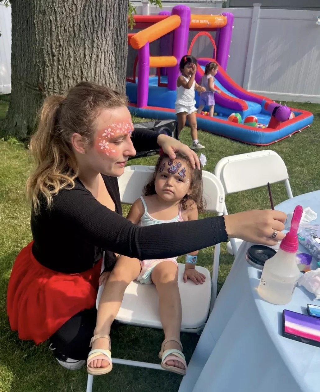 A woman with face paint of white flowers and a child with face paint of butterflies sitting at a table, while kids play in the background on an inflatable slide and bounce house in a backyard.