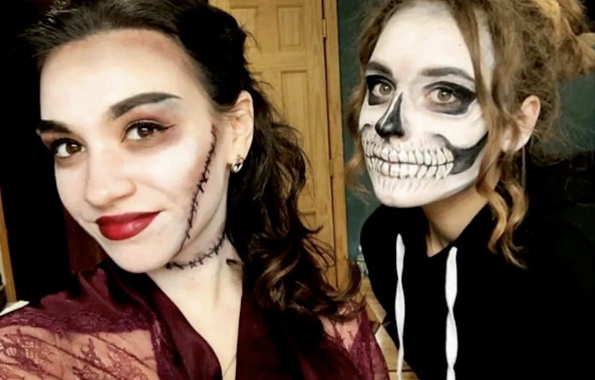 Two women in Halloween costumes; one woman has face makeup resembling a stitched scar with dark eye makeup and red lipstick, while the other woman has face makeup resembling a skull with black eye sockets and teeth, and wears a black hoodie.