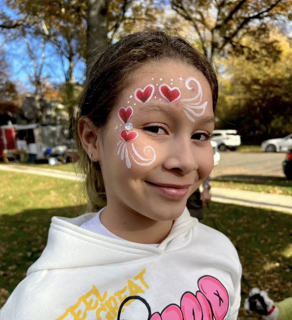 A young girl with face paint featuring red hearts, white swirls, and a gem on her cheek. She is smiling outdoors with fall foliage and parked cars in the background.