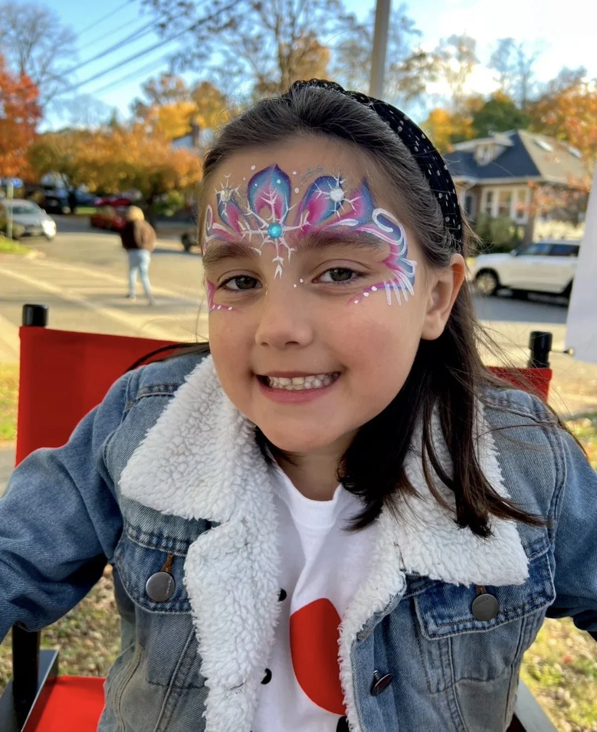 A young girl with face paint featuring a colorful, intricate butterfly design with stars and dots. She is smiling and wearing a denim jacket with a white, fluffy collar. The background shows a parking lot with autumn trees and houses.