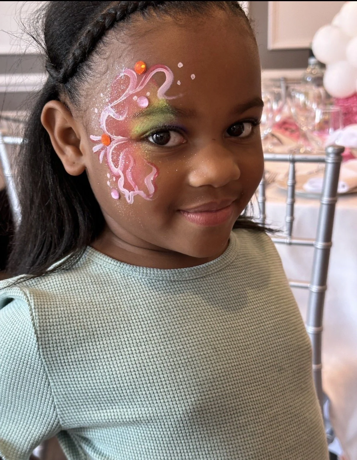 A young girl with face paint resembling a pink butterfly with lime green highlights and orange gems, smiling at a party.