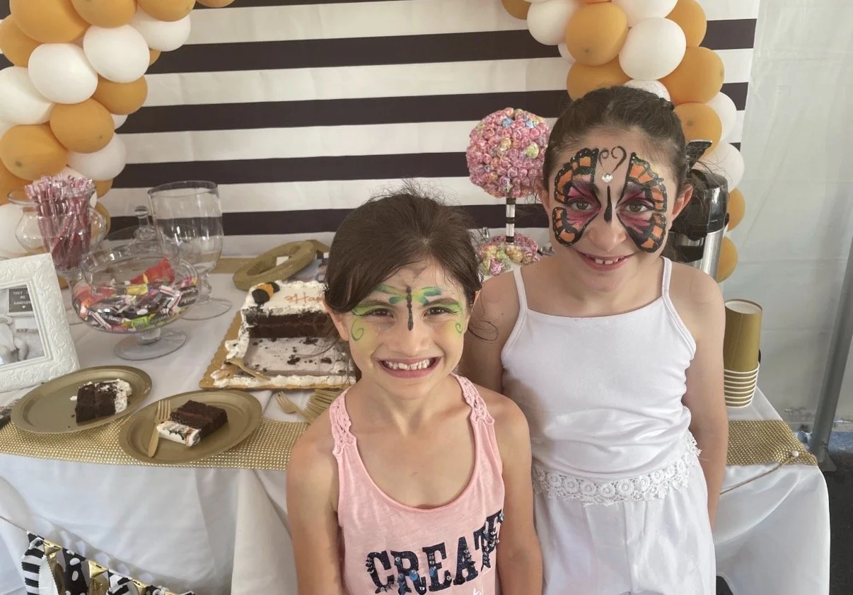Two young girls with butterfly face paint smiling at a birthday party with cake and decorations in the background.
