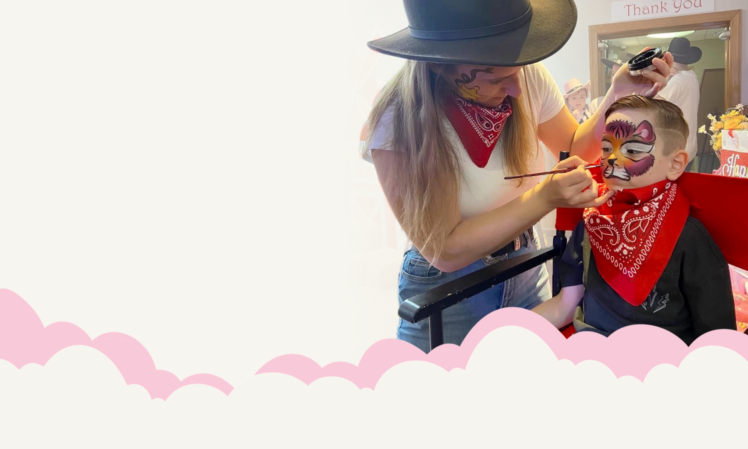 A woman with a cowboy hat and red bandana painting a tiger face on a young boy's face during a festive event.