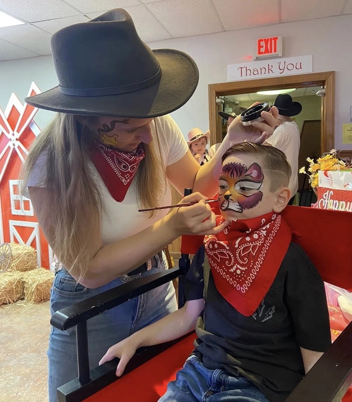 A woman is painting a tiger face on a young boy's face during a face painting activity. The boy is seated at a table and wearing a red bandana around his neck. The woman is wearing a large black cowboy hat and a red bandana around her neck. The backg