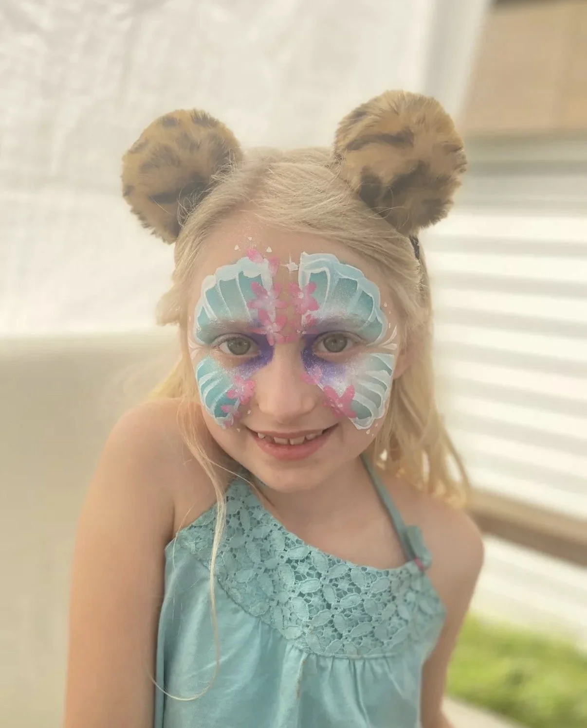 Young girl with painted butterfly face art, wearing a blue dress, and lion ears headband, smiling indoors with a blurred background.