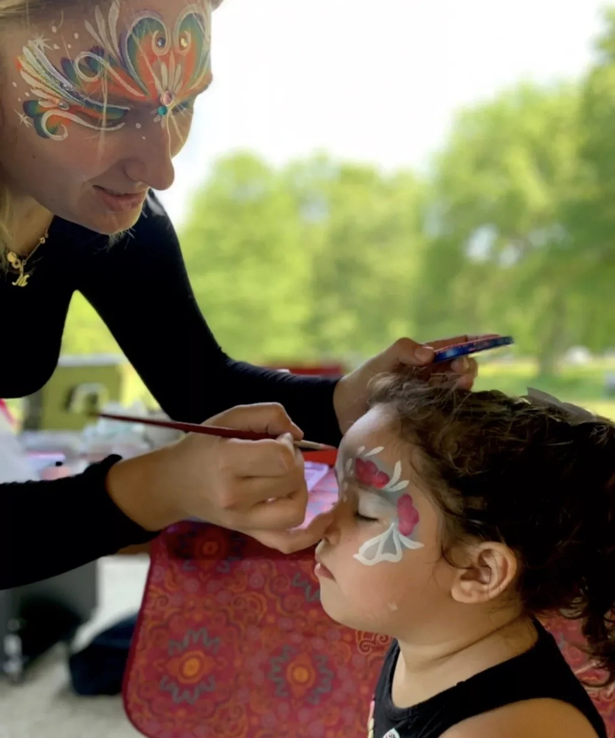 A woman painting a butterfly face art design on a young girl's face. The woman's face is painted with colorful swirls and designs that resemble a butterfly. The girl is sitting with eyes closed, wearing a dark tank top, and has curly hair. The backgr