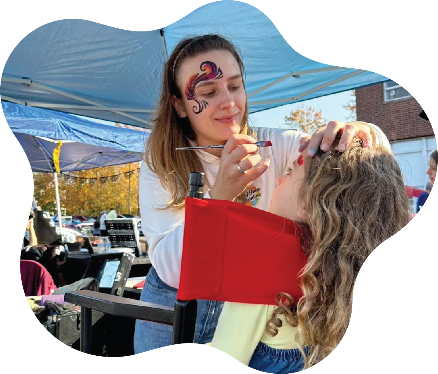 A woman with face paint in the shape of a colorful flame painting a young girl with curly hair on her forehead at an outdoor event under a blue canopy.
