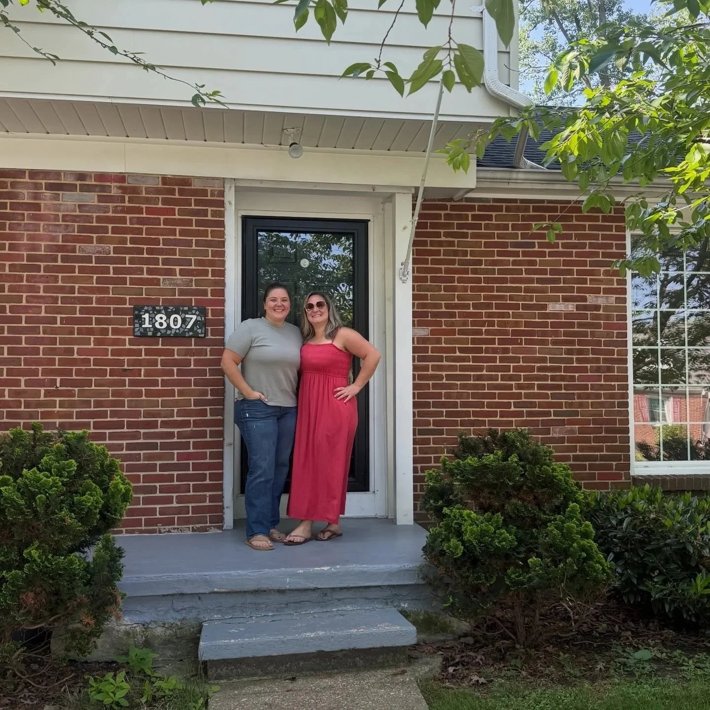Two women standing on the front porch of a brick house, smiling at the camera. One woman is wearing a grey t-shirt and jeans, the other is in a red maxi dress and wearing sunglasses. The house has a black front door and a house number sign that reads "1807". There are bushes and greenery in front of the porch.
