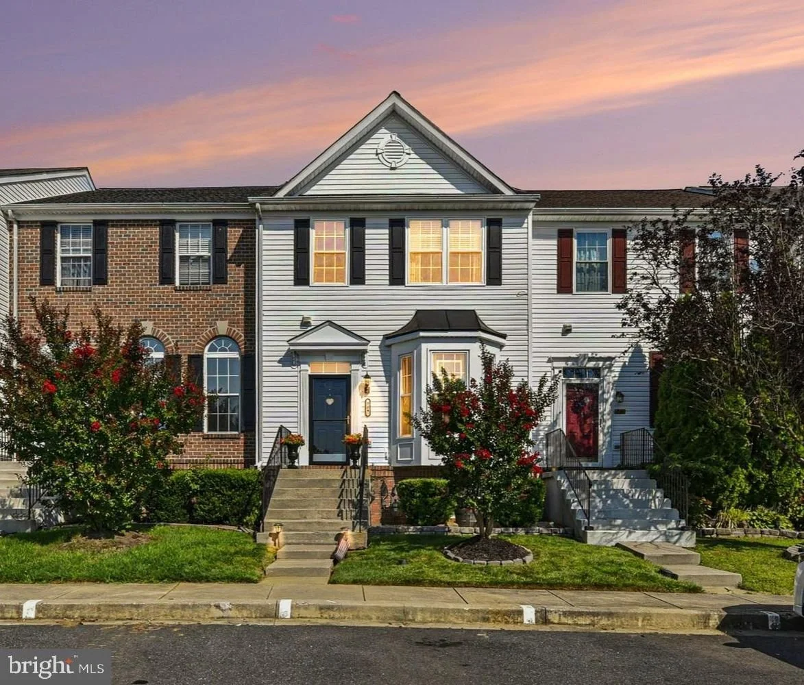 Front view of a two-story townhouse with white siding, black shutters, brick accents, and a small entrance porch, surrounded by landscaping and trees, under a colorful sunset sky.