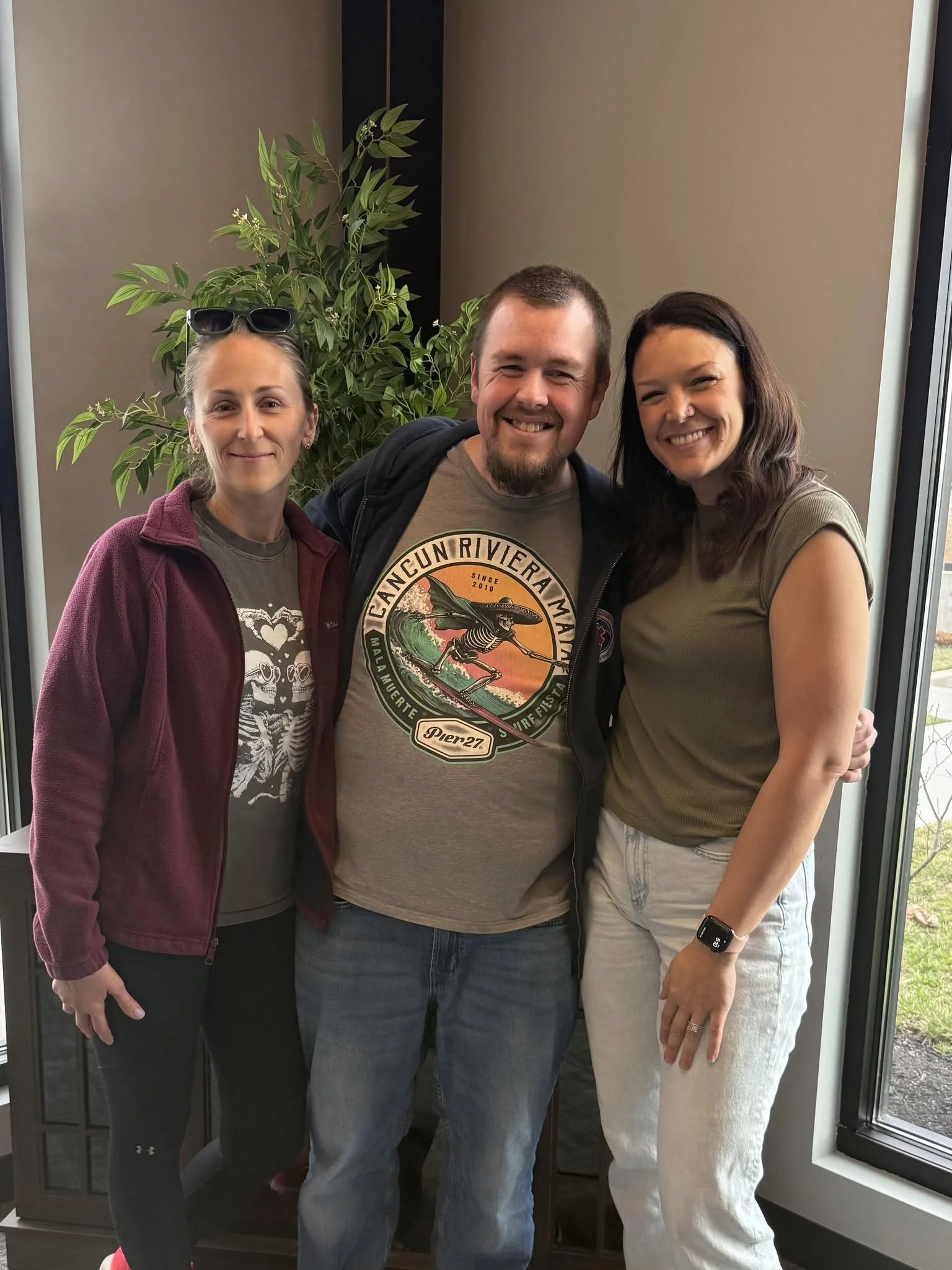 Three people standing closely together indoors near a window, smiling at the camera, with a green plant in the background.
